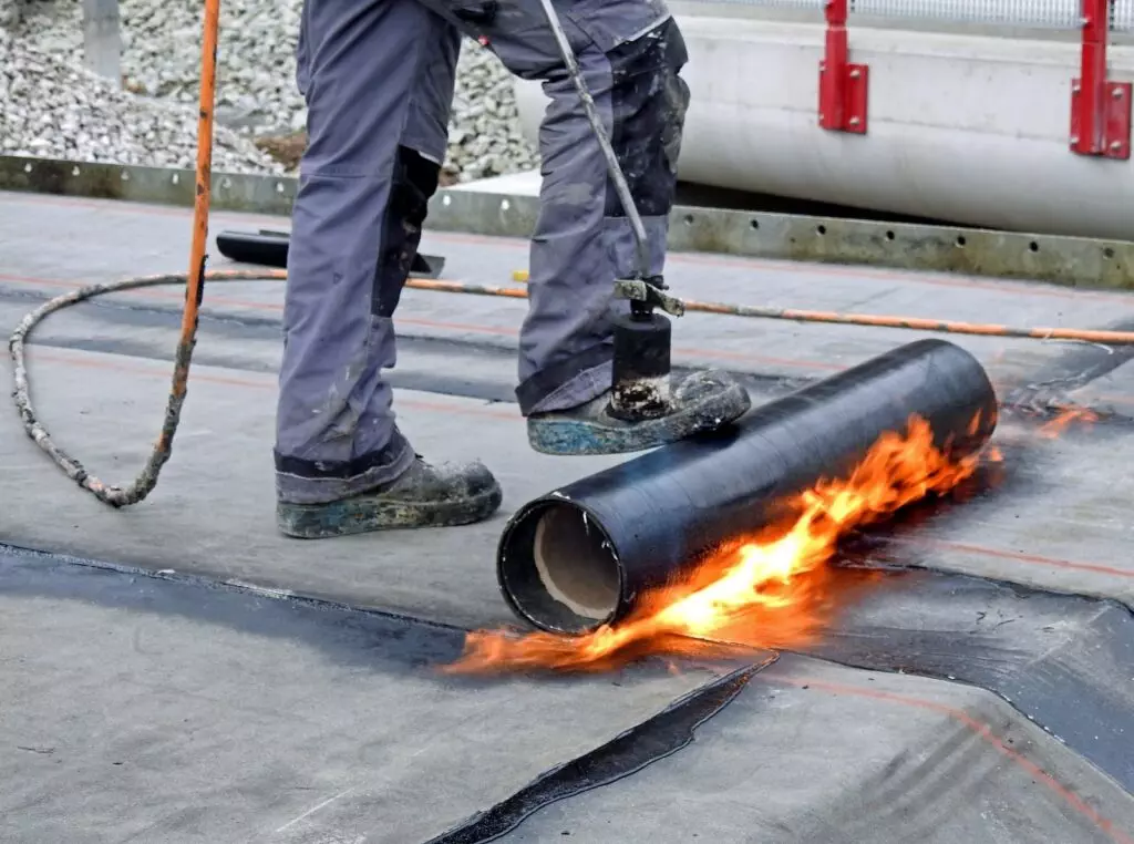 modified bitumen are rolled out onto the roof, and a roofing professional uses a hand-held propane torch to heat the material and adhere it to the surface